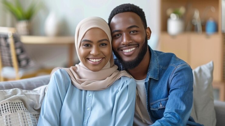 Une femme en hijab et un homme, souriants, assis côte à côte dans un salon.
